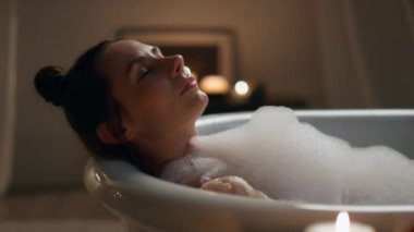 Bakımlı kız köpük banyosunu kapatıyordu. Kaygısız bir kadın akşam banyo yaparken gözlerini kapatıyor. Yumuşak cildi yıkayan bir kadın. İç mekândaki mumlarda bacaklarına dokunuyor. Model temizleme vücut dinlenme zamanı