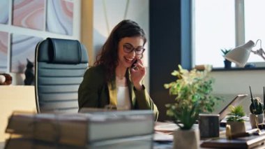 Neşeli, güzel kız ofis müdürü modern işyerinde oturarak akıllı telefondan konuşuyor. Gülümseyen iş kadını, yaratıcı şirket hizmetleri sunan müşteriyi arıyor. Kağıtlara bakan kız.