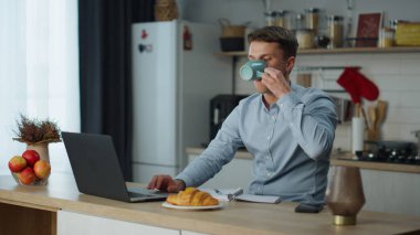 Modern, rahat bir mutfakta dizüstü bilgisayarla çalışan genç, çekici bir adam ve taze kahve. İnternette bilgi arayan genç bir iş adamı. Uzak iş yeri kavramı.