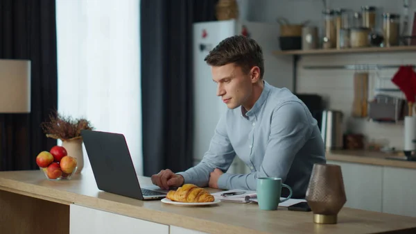 stock image Serious man searching internet working remotely on comfortable kitchen. Young guy freelancer looking at laptop screen checking email in morning. Busy worker using modern tech notebook device at home.