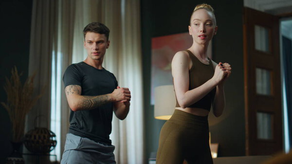 Attractive blond woman squatting with handsome man together in room. Young couple wearing sportswear exercising at home. Sporty family training legs strength. Fitness workout for pair indoors.