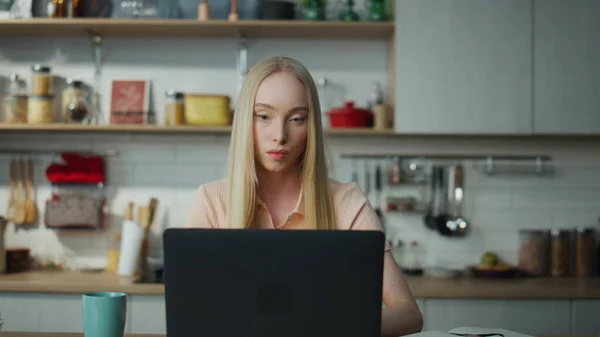 自信的金发女人坐在舒适的厨房特写镜头上 与笔记本电脑遥遥领先 专注的女商人在智能手机上浏览计算机上的信息 认真的女孩在网上工作 — 图库照片