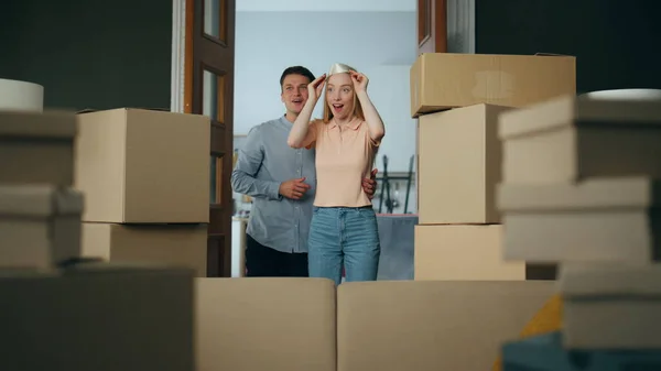 Excited newlyweds hugging moving at new apartment full of packed carton boxes. Young attractive woman surprising entering in modern living room. Cheerful family enjoy relocation in mortgage house.