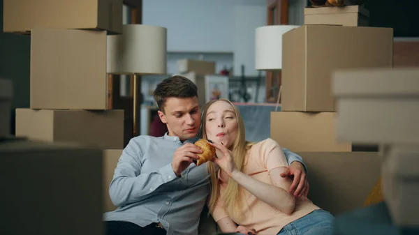 Happy family rent new house eating croissant on couch living room close up. Young couple relaxing hugging on sofa near cardboard boxes. Satisfied flat owners moving in modern mortgage apartment.