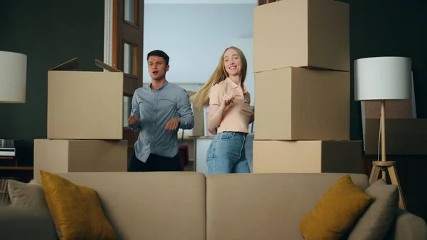 Positive couple dancing in new apartment between packed cardboard boxes. Young happy family making funny movements in own living room. Cheerful man rejoicing moving in modern flat with blond woman.