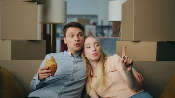 Relaxed young family breakfasting in new home sitting at comfortable couch between packed boxes close up. Happy cheerful couple eating croissant discussing interior new living room. Relocation concept