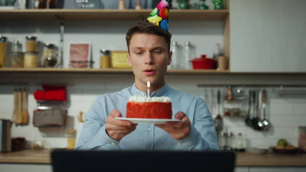 悲しい男ケーキろうそくパーティー帽子を着て吹いて単独でキッチンの近くに座っている 家族とのオンライン会議で誕生日デザートを祝う若い男を動揺させます 孤独を感じるハンサムな男 — ストック写真