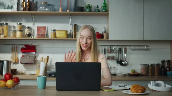 Happy beautiful girl chatting using web camera sitting at kitchen table alone. Attractive blond woman waving hand greeting friends on video call. Pretty lady smiling talking at zoom meeting on laptop.