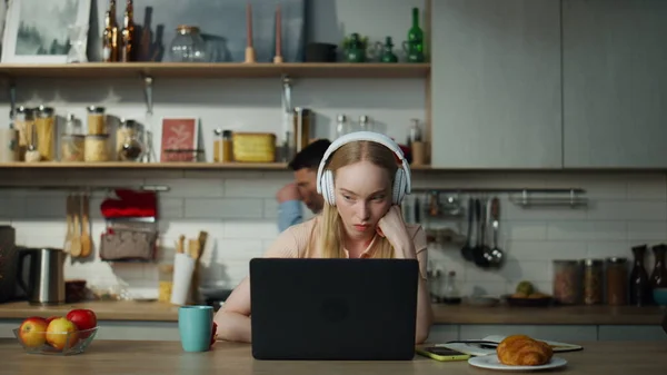 Dissatisfied girl programmer working laptop in headphones sitting at kitchen table close up. Focused girl freelancer upset by job failure looking screen. Husband dancing on backdrop distracting wife.