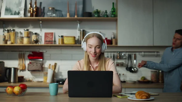 Pretty woman internet manager consulting online on video meeting wearing wireless headphones at home. Young cheerful guy dancing on backdrop. Attractive girl speaking on zoom conference using laptop.