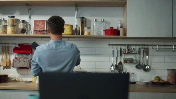 快乐的年轻人在厨房里跳舞 拿着长矛独自准备早餐 迷人而快乐的男人站在木制桌子上 在笔记本电脑旁接听视频电话 一个英俊的男人在现代公寓里做饭 — 图库照片