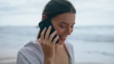 Gülümseyen neşeli kız kasvetli sahilde telefonu arıyor. Modern akıllı telefonla konuşan, boş deniz kıyısında yürüyen çekici mutlu bir kadının portresi. Güzel bayan doğada mobil iletişimden hoşlanır.