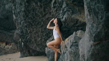 Taş kayaya yaslanmış sahilde poz veren seksi genç kadın. Deniz kıyısında beyaz mayo giyen çekici, şehvetli bir model. Uçurumun kenarındaki kumsalda duran muhteşem seksi kız. Güzellik konsepti.