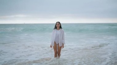 Seksi beyaz mayoyla deniz köpüğünde yürüyen dingin, güzel bir kadın. Uzun saçlı genç bayan kumlu okyanus plaj bulutlu gününe çıkıyor. Rahat çekici kız deniz kıyısında sakin ve kasvetli bir hafta sonunun tadını çıkar..