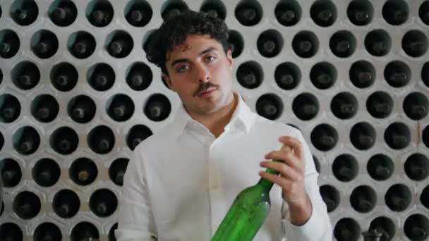 年轻的意大利葡萄酒鉴赏家拿着装有美酒的玻璃瓶走近镜头 背景下 站在葡萄酒厂酒窖旁的卷曲体力劳动者的画像 — 图库视频影像