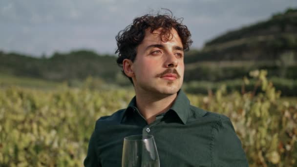 若い男のワイン醸造者は おいしい白ワインを閉じガラスのゴブレットを保持ブドウ畑を見ています 収穫とブドウのプランテーションを見て魅力的なイタリアのワインメーカー リラックスしたヴィンナーはワイナリービューをお楽しみください — ストック動画