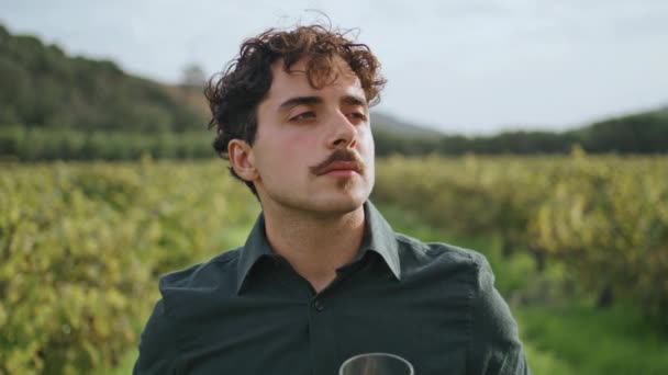 美しいブドウ畑に立つ若いイタリアのワインメーカーは ガラスの近くで絶妙な白ワインの匂いを楽しんでいます 中口ソムリエの肖像ブドウと谷で高級アルコールを味わう — ストック動画