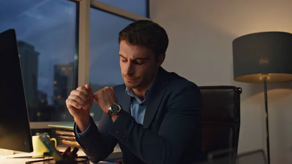 Pensive employee working night in office. Tired businessman checking hand watch staying late at workplace. Handsome successful executive analyzing financial report. Ceo planning strategy in lamp light