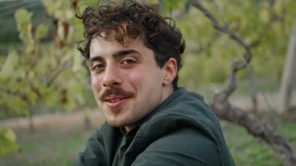 笑顔でカメラを探してブドウ畑に座って幸せな男の肖像画 プランテーションでブドウの茂みの下に座っている魅力的な口ひげの男が閉じます イタリアのワイン醸造家は収穫に満足してリラックス — ストック動画