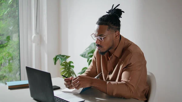 stock image Modern student learning remotely closeup. Stylish guy making notes at home. Focused freelancer watching webinar writing in notebook alone. African american man using laptop computer in light space 