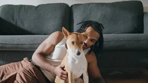 Happy man stroking dog at home closeup. Dreadlocks guy embracing domestic animal in stylish apartment alone zoom on. African american owner petting teasing his canine friend in living room interior
