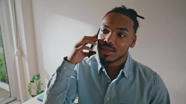 stock image Smiling guy talk smartphone at remote workplace. African american professional having conversation on cellphone in home office. Joyful man director call phone in light interior. Occupation concept 