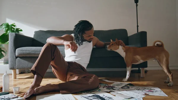 stock image Creative guy stroking dog at sofa room. African american man petting domestic animal sitting wooden floor. Positive artist relaxing after drawing modern sketches at home. Painter resting with hobby