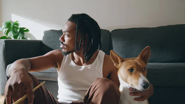 stock image Thoughtful guy embracing dog at sofa room closeup. African american man petting domestic animal sitting wooden floor. Brown skin artist relaxing after drawing sketches at home. Modern painter resting