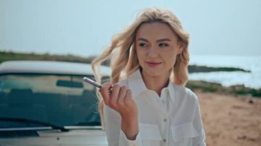 Güzel yaz plajında telefonla poz veren sevimli bir model. Yakından. Akıllı telefonlu çekici sarışın portresi güneşli deniz kıyısında duruyor ve retro arabaya yaslanıyor. Güzel kız turist görünümlü kamera..