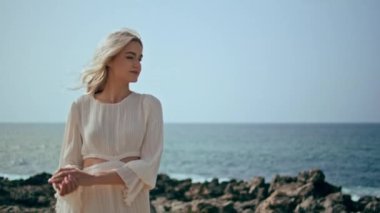 Romantik gezgin sahilde poz verip yaz güneşinin tadını çıkarıyor. Deniz kıyısında beyaz elbiseler giymiş güzel bir kadın. Çekici kaygısız sarışın okyanus kıyısında rahatlarken gülümsüyor..