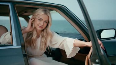 Güzel okyanus arkaplanındaki otomobil ön koltuğunda poz veren sevimli sarışın. Retro araba görünümlü kamerada poz veren kaygısız kadın. Çekici genç bayan gezgin inanılmaz deniz kıyısına araba yolculuğunun tadını çıkarıyor.