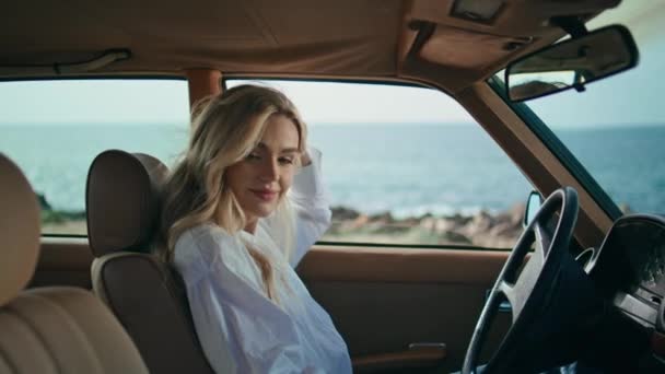 漂亮的金发姑娘坐在汽车掌舵上 紧紧抓住现代智能手机 迷人而轻松的模特儿 在美丽的阳光普照的海滨站在车内 微笑着看着相机 使用电话的妇女 — 图库视频影像
