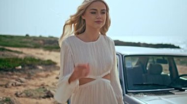 Sarışın kız arka planda eski arabasıyla deniz kıyısında yürüyor. Boş, güneşli okyanus kıyısında beyaz elbiseler içinde gezinen muhteşem genç bir kadın. Mutlu kaygısız bayan gezgin yaz yolculuğunun tadını çıkar..