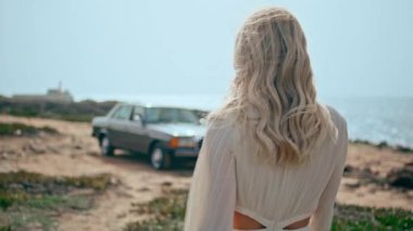Güzel bir yaz tatilinde sahilde yürüyen rahat bir kadın. Yakın plan. Deniz kıyısında duran eski moda arabaya giden mutlu güzel kız. Güzel sarışın model, güneşli okyanus kıyısında beyaz elbise giyerek poz veriyor..