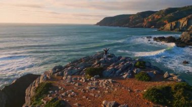 Okyanus kıyısı hava manzarasında silah sallayan bir adam. Bilinmeyen erkek, deniz doğasını izlerken kayalık kayalıklarda tek başına duruyor. Gezgin volkanik kayalarda el kaldırıyor. Süper yavaş çekim. Özgürlük kavramı