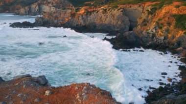 Fırtınalı köpüklü okyanusun üzerinde sinematik manzaralı kayalık bir sahil. Drone görüntüsü güneşli deniz havuzu güçlü deniz dalgaları tehlikeli volkanik kayalıklara yavaş çekimde çarpıyor. Güzel dalgalar taşlara sıçrıyor