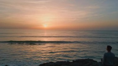 Karanlık uçurumların ardında gün batımını izleyen bir adam silueti. Panoramik şafak deniz manzarası drone görüntüsü. Sonsuz manzaralı okyanus ufku. Pastel pembe ufuk çizgisi. Portakal rengi günbatımı, sakin suyla yansıyor.