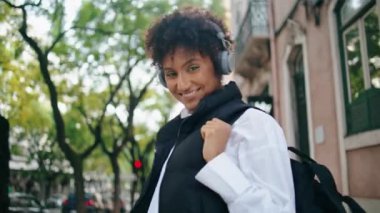 Modern kulaklıklar takan pozitif turist kız. Dışarıda poz verip kulaklıkla müzik dinleyen kaygısız Afrikalı Amerikalı kadın. Neşeli hippi bayan kasabada gezintiye çıkar..