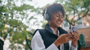 Neşeli Afro-Amerikalı bayan cep telefonuyla kablosuz kulaklıkla müzik dinliyor. Şehir caddesinde duran telefon uygulamasını kullanan çekici, mutlu bir kadın. Pozitif kız en sevdiği şarkıyı seviyor..
