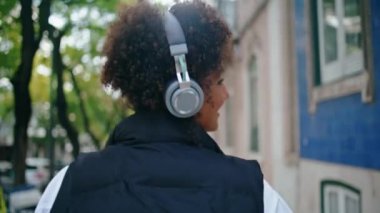 Şehir caddesinde yürüyen, kablosuz kulaklıklarla müzik dinleyen kaygısız bir kadın. Kulaklıklı mutlu Afro-Amerikalı kız dışarıda gülümseyerek kameraya dönüyor. Hanımefendi, sakin bir gezinti yapın..