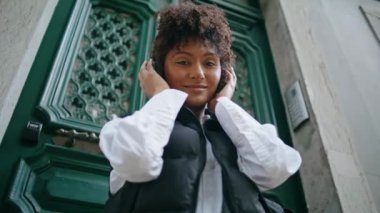 Rahat Afro-Amerikan kadın kulaklıklı klasik kapı önünde dikiliyor. Kablosuz kulaklık takıp kameraya gülümseyen mutlu çekici kız. Dışarıda müzik dinleyen bir bayan..
