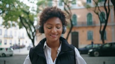 Rahat Afro-Amerikan kadınları, kablosuz kulaklıklarla şehrin caddesini kapatan şarkılardan hoşlanırlar. Modern kulaklıklarla müzik dinleyen mutlu gülen kız. Güzel bayan ritmik olarak hareket ediyor..