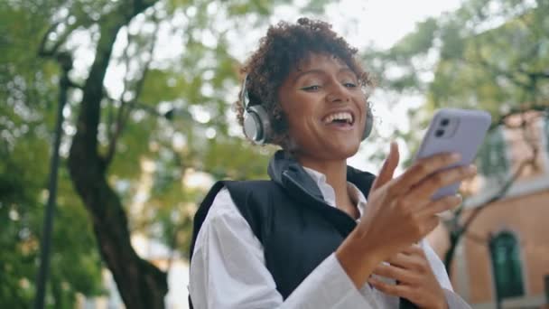 快乐的微笑的女人站在城市街道的近旁 在无线耳机上打开最喜爱的音乐 兴奋的非洲裔美国女孩喜欢智能手机上的移动应用 卷曲的女士沿着有节奏的歌曲跳舞 — 图库视频影像