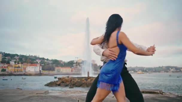 曇り空の桟橋でラテンダンスを行う熱い美しいパートナー プロのダンサーの近くに魅惑的に腰を動かす青いドレスのセクシーなブルネットの女性 情熱的な若いですカップルダンスで海岸スローモーション — ストック動画