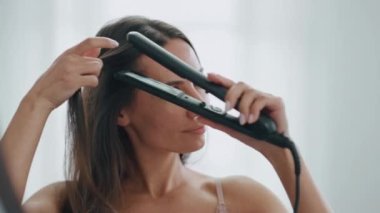 Beyaz banyo odasında saç ütüsü kullanan mutlu bir kız. Gülümseyen uzun saçlı kadın sabah saç stili yapıyor. Duşta esmer Chevelure aynasını düzelten güzel bayan. Ev rutini