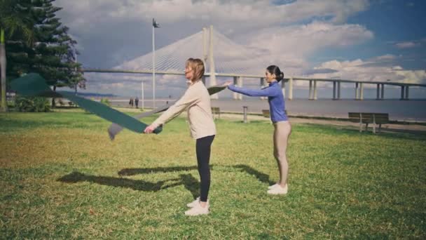 瑜伽女孩把地毯放在草地上 两名健康的女性准备在新鲜空气中锻炼 活跃的身材苗条的女士在绿园上普拉提训练班 有动力的运动员参加体育项目 健康的人 — 图库视频影像