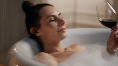 Romantik bir bayan küveti soğutuyor ve kırmızı şarap içiyor. Gülümseyen kadın sıcak köpüklü suda alkollü içecekten hoşlanır. Rahat ol. Bakımlı iyimser kız geceyi mum banyosunda geçiriyor.