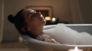 Bakımlı kız köpük banyosunu kapatıyordu. Kaygısız bir kadın akşam banyo yaparken gözlerini kapatıyor. Yumuşak cildi yıkayan bir kadın. İç mekândaki mumlarda bacaklarına dokunuyor. Model temizleme vücut dinlenme zamanı