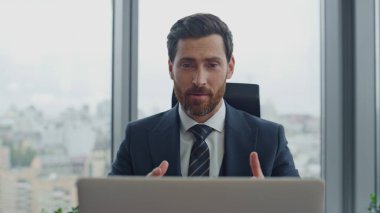Başarılı sakallı yöneticinin laptopta video görüşmesi var. Ofis masasını kapatıyorlar. Yakışıklı girişimci iş ekibiyle çevrimiçi toplantıda konuşuyor. Gülümseyen patron video çağrısının keyfini çıkar..