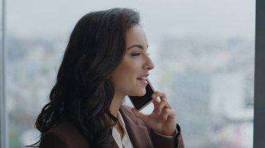 Gülümseyen çekici iş kadını iş yerinde telefonda konuşuyor. Panoramik pencerenin önünde duruyor. Esmer kız yöneticinin portresi. Modern akıllı telefon kullanarak arkadaşlarıyla gülüyor..